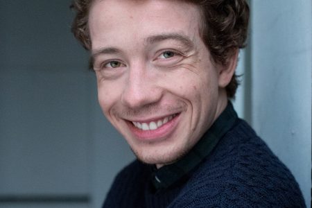 Ein junger Mann mit lockigen braunen Haaren und einem blauen Pullover lächelt in einer schwach beleuchteten Innenumgebung in die Kamera.