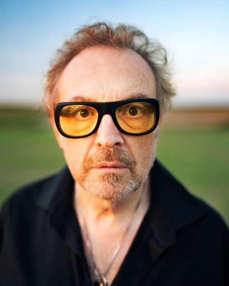Ein Mann mit zerzaustem Haar und Bart, der eine große gelbe Brille und ein schwarzes Hemd trägt, steht im Freien mit einem verschwommenen grünen Feld im Hintergrund.