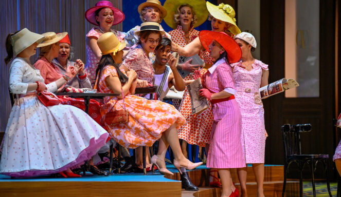 Eine Gruppe von Frauen in bunten Vintage-Kleidern und Hüten sitzt und steht lachend und schwatzend um einen kleinen Tisch auf der Bühne.
