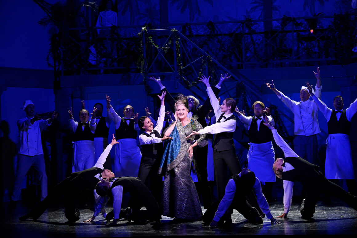 Eine Frau in einem eleganten Kleid singt in der Mitte der Bühne, umgeben von Darstellern in schwarz-weißen Kostümen mit ausgestreckten Armen in einer dramatischen Musicalszene.