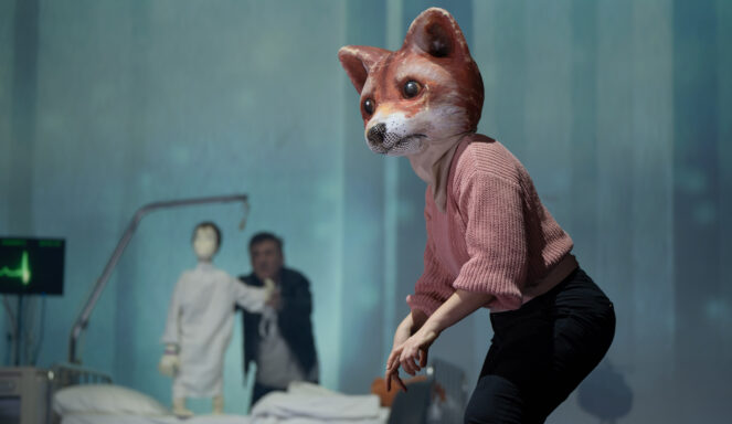 Eine Person mit einer großen Fuchsmaske und einem rosafarbenen Pullover steht im Vordergrund, während im Hintergrund ein Krankenhausbett und zwei Figuren verschwommen zu sehen sind.