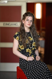 Eine junge Frau mit langen braunen Haaren trägt ein Kleid mit Sonnenblumenmuster und Tupfen, eine Perlenkette und Armbänder. Sie sitzt in einem Haus in der Nähe einer Tür mit der Aufschrift "EINGANG".