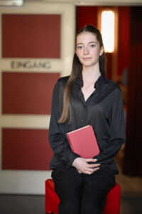 Eine junge Frau mit langen braunen Haaren und schwarzem Hemd sitzt mit einem roten Buch in der Hand vor einer Tür mit der Aufschrift "EINGANG" im Hintergrund.