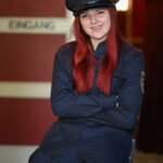 Eine Person mit langen roten Haaren, gekleidet in eine dunkle Polizeiuniform mit Hut, sitzt auf einem roten Stuhl und lächelt mit verschränkten Armen.