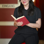 Eine Frau mit Brille und dunklem Haar sitzt auf einem roten Hocker, lächelt und hält ein aufgeschlagenes Buch in der Hand, im Hintergrund ein Schild mit der Aufschrift "EINGANG".