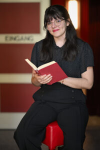 Eine Frau mit Brille und dunklem Haar sitzt auf einem roten Hocker, lächelt und hält ein aufgeschlagenes Buch in der Hand, im Hintergrund ein Schild mit der Aufschrift "EINGANG".