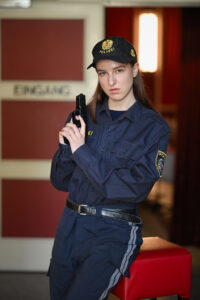 Ein seriöser Polizeibeamter in Uniform und mit Mütze hält eine Pistole in der Hand und steht in einem Innenraum in der Nähe einer Tür mit der Aufschrift "Eingang" (Deutsch für "Eingang").