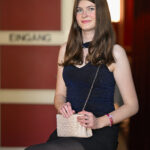 Eine junge Frau in einem blauen Kleid sitzt auf einem roten Hocker und hält eine kleine Handtasche in der Hand. Im Hintergrund befindet sich eine Tür mit der Aufschrift "Eingang".