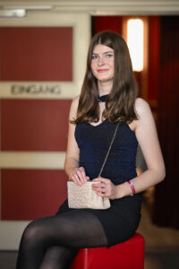 Eine junge Frau in einem blauen Kleid sitzt auf einem roten Hocker und hält eine kleine Handtasche in der Hand. Im Hintergrund befindet sich eine Tür mit der Aufschrift "Eingang".