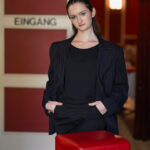 Eine Frau in einem dunklen Anzug steht mit den Händen in den Taschen neben einem roten Hocker vor einer Tür mit der Aufschrift "Eingang" und roten Wänden.
