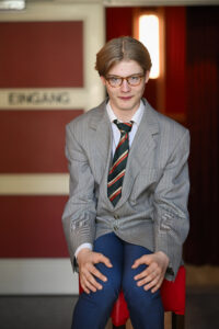 Ein junger Mann mit Brille und gestreifter Krawatte sitzt lächelnd auf einem roten Stuhl, im Hintergrund steht ein Schild mit der Aufschrift "Eingang".