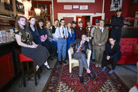 In einer Bar mit roten Wänden und einem gemusterten Teppich posiert eine Gruppe von Menschen in Vintage-Kostümen, einige sitzend, andere stehend.