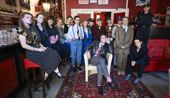 In einer Bar mit roten Wänden und einem gemusterten Teppich posiert eine Gruppe von Menschen in Vintage-Kostümen, einige sitzend, andere stehend.