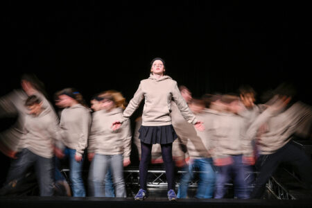 Eine Person steht still auf der Bühne vor einer verschwommenen Menge, die Arme ausgestreckt, mit einem beigen Kapuzenpullover, einem schwarzen Rock und blauen Schuhen.