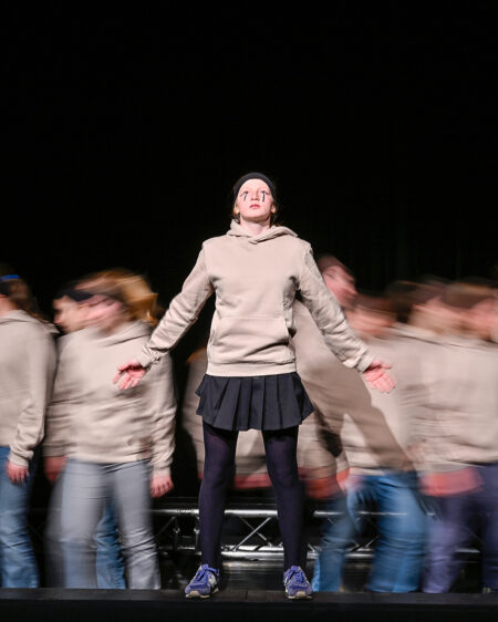 Eine Person steht still auf der Bühne vor einer verschwommenen Menge, die Arme ausgestreckt, mit einem beigen Kapuzenpullover, einem schwarzen Rock und blauen Schuhen.