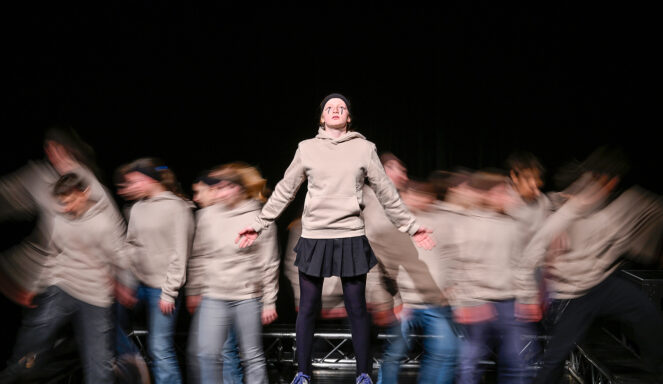 Eine Person steht still auf der Bühne vor einer verschwommenen Menge, die Arme ausgestreckt, mit einem beigen Kapuzenpullover, einem schwarzen Rock und blauen Schuhen.