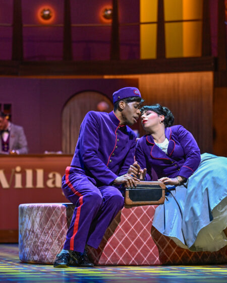 Zwei Schauspieler in lila Uniformen sitzen dicht beieinander auf der Bühne, kurz vor einem Kuss, mit Koffern neben ihnen und einer alten Hotelrezeption im Hintergrund.