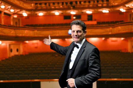 Ein Mann im Smoking steht auf der Bühne, lächelt und gestikuliert in Richtung eines leeren, reich verzierten Theaters mit Sitzreihen und Balkonen.