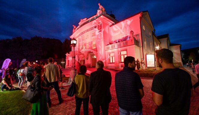 Menschen versammeln sich nachts vor einem beleuchteten Gebäude, um eine Lichtprojektion auf dessen Fassade zu sehen. Die Szene ist lebendig und pulsierend.