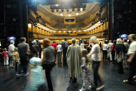 Eine Gruppe von Menschen steht und geht auf einer Theaterbühne mit Blick auf die Zuschauerplätze in einem hell erleuchteten, reich verzierten Zuschauerraum.