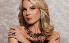 Blonde Frau, die ein schwarzes Oberteil mit Federn, eine bunte Edelsteinkette, einen bunten Ring und Ohrringe trägt und mit verschränkten Armen vor einem gemusterten Hintergrund posiert.