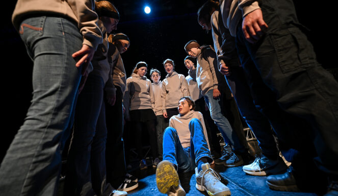 Eine Gruppe von Menschen in legerer Kleidung steht im Kreis und blickt auf eine Person, die unter dramatischer Bühnenbeleuchtung auf dem Boden sitzt.