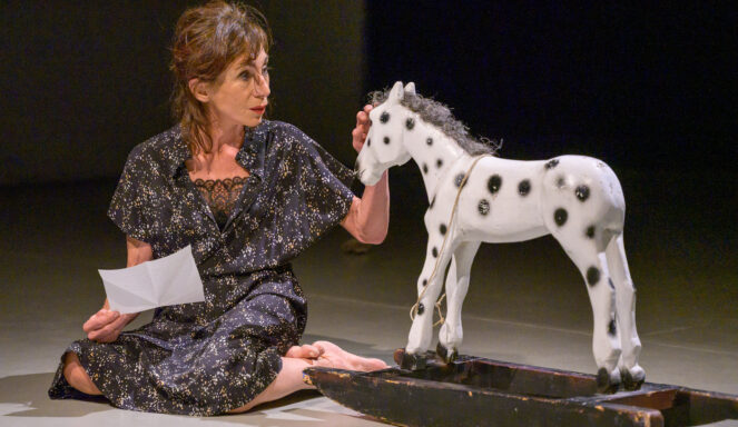 Eine Frau in einem gemusterten Kleid sitzt barfuß auf dem Boden, hält eine Zeitung in der Hand und berührt ein weißes, gepunktetes Schaukelpferd.