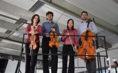 Vier Musiker stehen auf einer Metallplattform und halten jeweils ein Instrument in der Hand: zwei Geigen, eine Flöte und ein Cello, in einem modernen Gebäude.