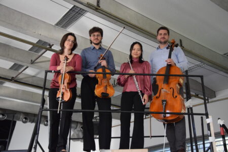 Vier Musiker stehen auf einer Metallplattform und halten jeweils ein Instrument in der Hand: zwei Geigen, eine Flöte und ein Cello, in einem modernen Gebäude.