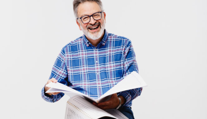 Lächelnder älterer Mann mit Brille und blau kariertem Hemd, der vor einem schlichten weißen Hintergrund geöffnete Notenblätter oder Dokumente hält.