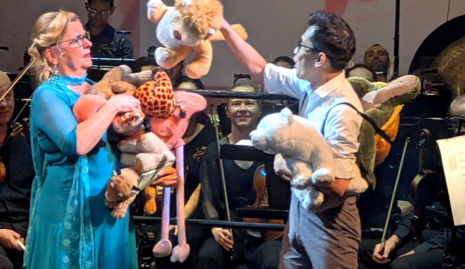 Zwei Personen auf der Bühne halten ausgestopfte Tiere, die spielerisch miteinander interagieren, während die Orchestermusiker im Hintergrund sitzen und zusehen.