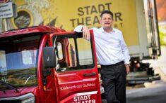 Ein Mann mit weißem Hemd lehnt sich an ein rotes Stadttheater-Fahrzeug, während im Hintergrund ein gelber Stadttheater-LKW zu sehen ist.