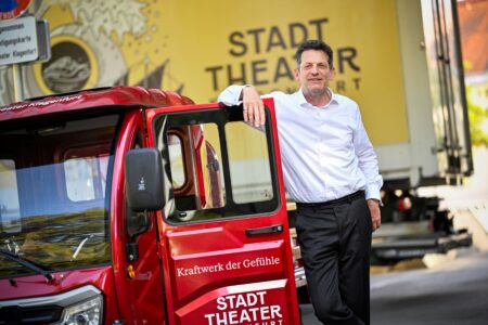 Ein Mann mit weißem Hemd lehnt sich an ein rotes Stadttheater-Fahrzeug, während im Hintergrund ein gelber Stadttheater-LKW zu sehen ist.