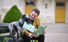 Ein Mann in einer hellbraunen Jacke und ein Hund sitzen auf einer Bank und scheinen gemeinsam grüne Broschüren vor einem Gebäude mit gelben Türen zu lesen.