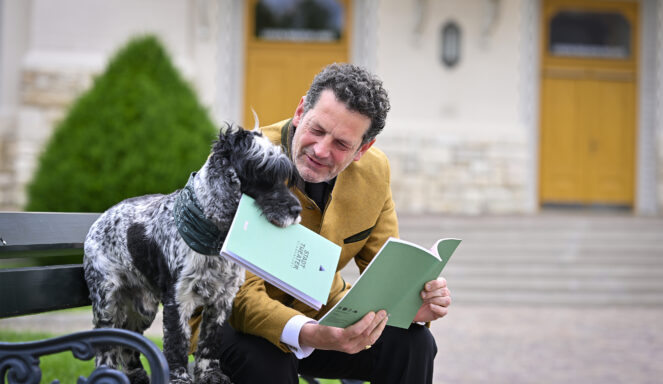 Ein Mann in einer hellbraunen Jacke und ein Hund sitzen auf einer Bank und scheinen gemeinsam grüne Broschüren vor einem Gebäude mit gelben Türen zu lesen.