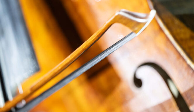 Nahaufnahme eines Cellobogens, der über die Cellosaiten gezogen wird, wobei die hölzerne Oberfläche des Instruments und das F-Loch im Hintergrund zu sehen sind.