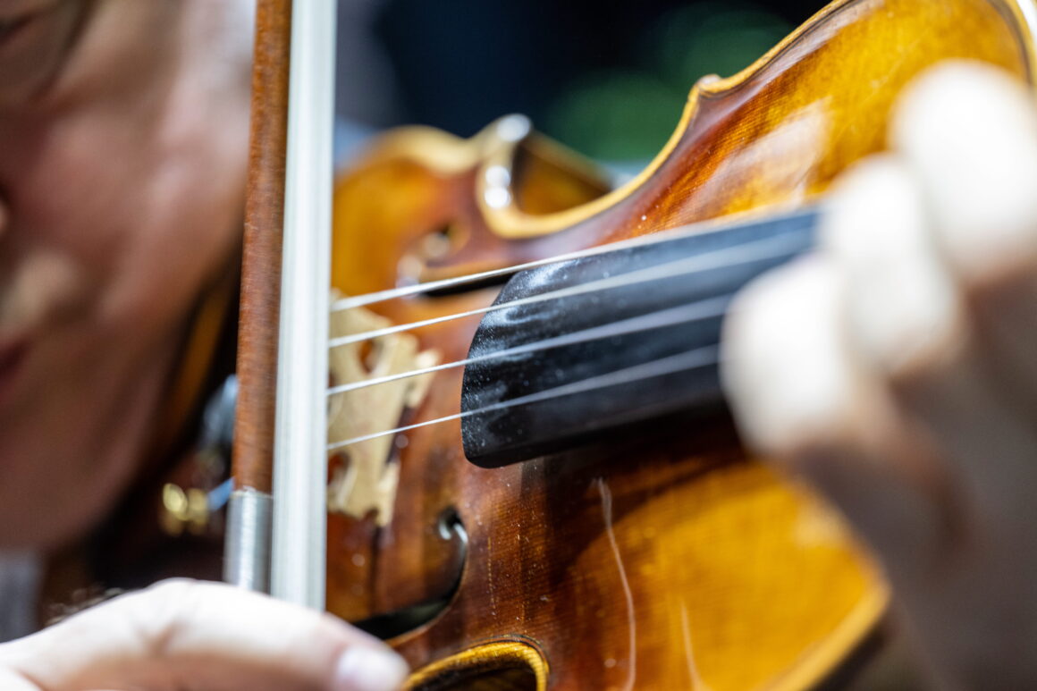 Nahaufnahme einer Geige spielenden Person, wobei der Schwerpunkt auf der Hand liegt, die die Saiten anschlägt, und dem Bogen, der sich über das Instrument bewegt.