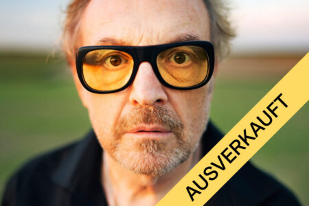 Nahaufnahme eines Mannes, der eine gelb getönte Brille trägt, mit einem gelben, diagonal verlaufenden Banner, auf dem „AUSVERKAUFT“ steht.