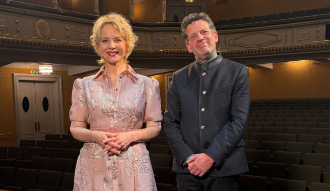 Eine Frau in einem hellrosa Kleid und ein Mann in einem dunklen Anzug stehen lächelnd zusammen in einem leeren Theater mit Sitzreihen hinter ihnen.