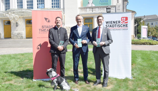 Drei Männer in Anzügen stehen auf einer Wiese und halten Broschüren in der Hand, flankiert von Bannern; vor ihnen sitzt an einem sonnigen Tag ein kleiner schwarz-weißer Hund.