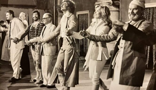 Auf einem alten Schwarz-Weiß-Foto ist zu sehen, wie eine Gruppe kostümierter Darsteller nach einer Theateraufführung auf der Bühne lächelnd Händchen hält und sich verbeugt.