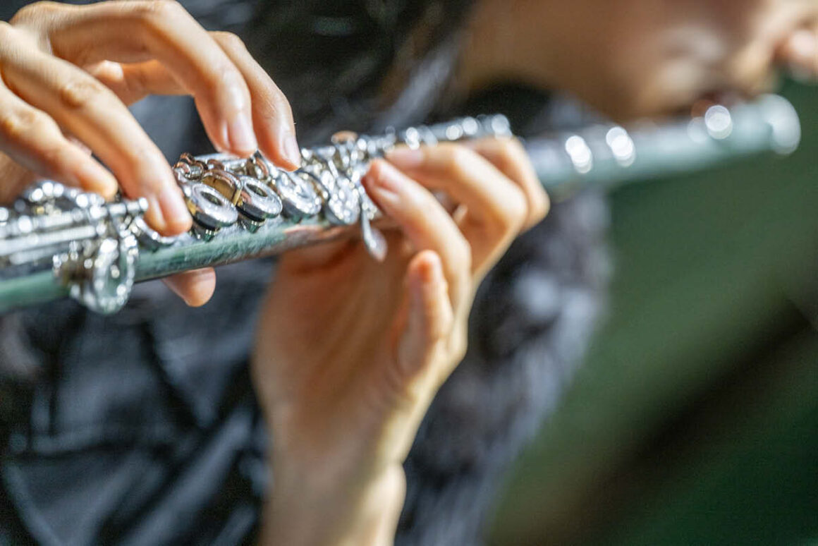 Nahaufnahme der Hände einer Person, die auf einer silbernen Flöte spielt, mit unscharfem Hintergrund und Fokus auf die Klappen des Instruments.