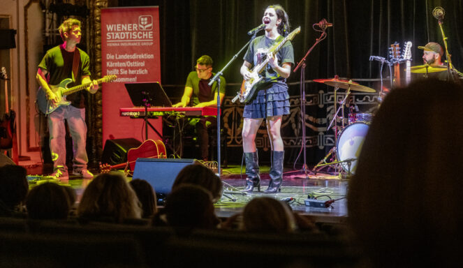 Eine Band tritt auf der Bühne auf; eine Frau singt und spielt Gitarre, begleitet von Musikern am Keyboard, am Bass und am Schlagzeug, während das Publikum zuschaut.