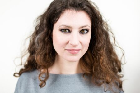 Eine Frau mit langen, lockigen braunen Haaren, die ein graues Oberteil trägt und vor einem schlichten weißen Hintergrund mit einem leichten Lächeln in die Kamera blickt.