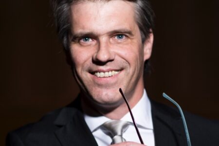 Ein lächelnder Mann in Anzug und Krawatte, der eine Brille in der Hand hält.